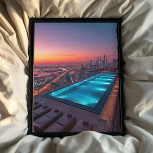 May include: A framed photograph featuring a vibrant sunset over a city skyline. The image showcases a rooftop pool with illuminated blue water, lounge chairs, and a view of skyscrapers. The frame is black, set against a backdrop of a white bed sheet.