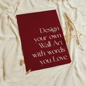 Może przedstawiać: Prostokątny ciemnoczerwony wydruk z białym napisem: Design your own Wall Art with words you Love. Wydruk umieszczono na teksturowanej powierzchni z tkaniny w kolorze złamanej bieli obok małego pęczka suszonych beżowych łodyg pszenicy.