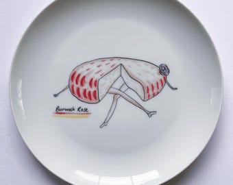 May include: A white ceramic plate with a hand-painted illustration of a red and white cheese wheel with legs and a flower on its head. The cheese is labeled "Burwash Rose".