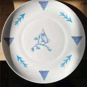 Könnte beinhalten: Weiße Keramikplatte mit einem blauen und weißen Eulen-Design. Die Eule trägt einen blauen Hut und sitzt auf einem Ast. Der Teller ist mit blauen Dreiecken am Rand verziert.