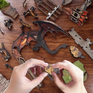May include: A wooden dragon puzzle with a detailed design, featuring a dragon with red and black wings. The puzzle pieces are scattered on a wooden surface, with hands assembling a piece. Other pieces include castle structures and trees.