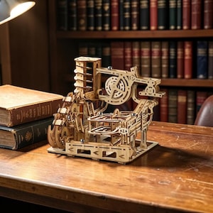 Op de afbeelding: Een houten mechanisch model met tandwielen en rails. Het model is gemaakt van lichtgekleurd hout en heeft ingewikkelde uitgesneden ontwerpen. Het staat op een houten oppervlak, met boeken en een boekenkast op de achtergrond.