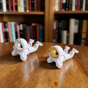 May include: Two white astronaut figurines in a prone position, resting on their stomachs. One astronaut features a reflective silver helmet visor, while the other astronaut features a reflective gold helmet visor. Both figures have detailed suits.