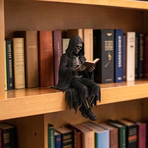 May include: A figurine of a skeleton wearing a black hooded robe and sitting on the edge of a wooden shelf. The skeleton is holding an open book in its hands, with its ribcage visible beneath the robe. It wears dark shoes and has a skull head.