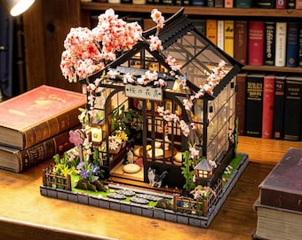 Puede incluir: Casa japonesa en miniatura con techo negro, paredes de cristal y un pequeño jardín. La casa está adornada con flores de cerezo rosas y un pequeño letrero con texto japonés. Un gato está dentro.