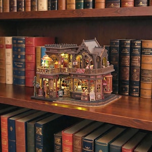 Pu&ograve; includere: Modello in miniatura dettagliato di una libreria e caffetteria, con calda illuminazione interna. La struttura a due piani presenta intricati dettagli architettonici, tra cui balconi e tende da sole. Il modello &egrave; esposto su una mensola di legno circondata da libri antichi.