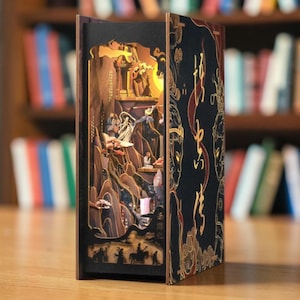 May include: A decorative wooden book with a detailed, layered interior scene illuminated by a warm light. The open book reveals a landscape with figures, mountains, and a pagoda. The cover is black with gold and red accents.