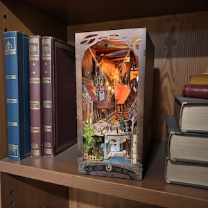 May include: A wooden Lord of the Rings-themed light box featuring a detailed miniature town scene with buildings, a bridge, and figures. The box has a warm, orange glow from the interior lighting. The text "THE LORD OF THE RINGS" is visible on the base.