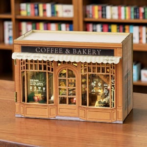 May include: A miniature model of a coffee shop and bakery with the text "COFFEE & BAKERY" on a sign above the awning. The shop has large windows displaying interior details and is made of light brown wood. The model is set against a backdrop of bookshelves.