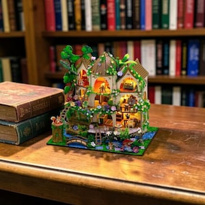 May include: A miniature fairy house with intricate details, including a bridge, flowers, and a tree. The house is illuminated, showcasing interior furnishings. It sits on a wooden surface with antique books in the foreground, against a backdrop of a bookshelf.