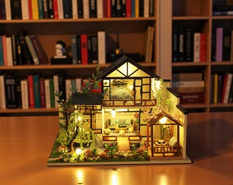 May include: Miniature wooden house with interior lighting. The two-story structure features a black roof, a balcony, and a small garden with miniature plants. The interior is lit, showcasing furniture and decor. The house is set on a wooden table.