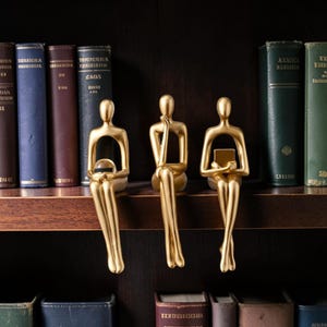 Pode incluir: Três figuras humanas abstratas de cor dourada sentadas numa prateleira. A figura da esquerda segura uma esfera transparente, a figura central apoia uma mão no queixo e a figura da direita segura um pequeno objeto retangular sem rosto.