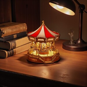May include: A miniature carousel music box with a red and white striped canopy and gold accents. The carousel features illuminated horses and is set on a wooden surface, with a warm glow emanating from within. A vintage lamp and books are in the background.