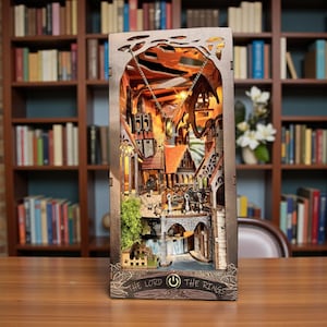 May include: A wooden Lord of the Rings-themed book nook with intricate laser-cut details. The diorama features a fantasy town scene with buildings, bridges, and a dragon. The text "THE LORD OF THE RINGS" is visible on the base. The scene is illuminated with warm lighting.
