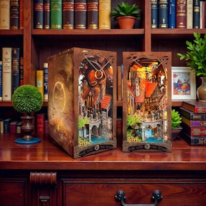 May include: Two intricately designed wooden book nooks depicting scenes from "The Lord of the Rings." Each features a detailed miniature cityscape with bridges, buildings, and a glowing ring. The nooks are brown with warm lighting, creating a cozy, fantasy-themed display.
