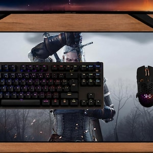 May include: A rectangular desk mat featuring a graphic print of a character from The Witcher 3 Wild Hunt. The mat is black with a gray forest background and includes a black mechanical keyboard and a black gaming mouse placed on its surface.