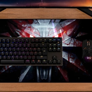 May include: A rectangular black desk mat featuring a large, stylised wolf head emblem in white with red accents and glowing red eyes. The mat is positioned under a black mechanical keyboard and a black computer mouse, providing a wide surface area.