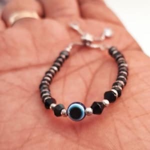 May include: Adjustable bracelet featuring a central blue and white evil eye bead, flanked by black faceted beads and smaller silver-toned beads. The bracelet is strung on a silver-toned chain.