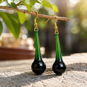 May include: A pair of drop earrings featuring elongated, teardrop-shaped glass pendants that transition from a deep forest green at the top to a solid black at the base. The pendants are suspended from gold-toned metal fishhook ear wires.