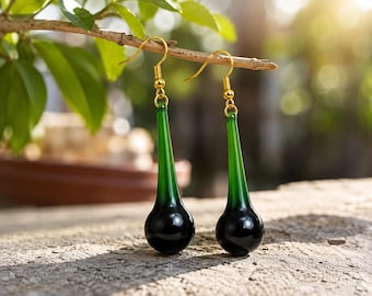 May include: A pair of drop earrings featuring elongated, teardrop-shaped glass pendants that transition from a deep forest green at the top to a solid black at the base. The pendants are suspended from gold-toned metal fishhook ear wires.