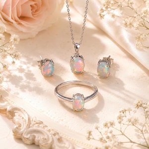 May include: A jewelry set featuring a silver necklace, ring, and earrings, each with an oval opal gemstone. The opal stones display a mix of colors, including pink, blue, and yellow. The set is displayed on a cream-colored surface with floral accents.