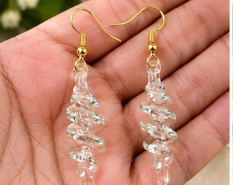 May include: A pair of clear glass earrings featuring a twisted spiral design. Each earring hangs from a gold-toned metal hook. The glass elements are transparent and have a smooth, reflective surface, creating a distinct corkscrew shape.