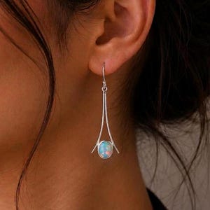 May include: Silver-toned drop earrings with a teardrop design. Each earring features a long, slender bar that widens to cradle a round, opalescent stone. The stones display a mix of pale blue, pink, and white hues.