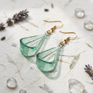 May include: A pair of drop earrings featuring translucent, light green, teardrop-shaped glass pendants with a textured surface. The earrings are attached to gold-toned metal fishhook wires, providing a simple and elegant design for these glass accessories.