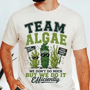 Peut inclure: Un t-shirt couleur crème avec un motif portant le texte "TEAM ALGAE" et "WE DON'T DO MUCH BUT WE DO IT EFFICIENTLY." Le design présente trois personnages d'algues verts, l'un tenant une pancarte "MICRO BUT MIGHTY" et l'autre "OXYGEN PRODUCING LEGENDS."