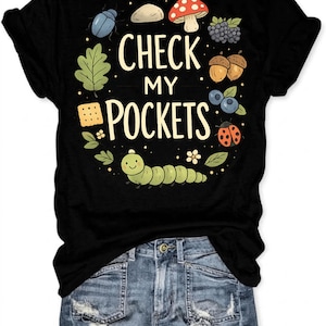 May include: Black t-shirt with a whimsical design featuring the text "CHECK MY POCKETS" surrounded by illustrations of a beetle, mushroom, berries, acorns, and a smiling caterpillar. The design is in a circular arrangement.