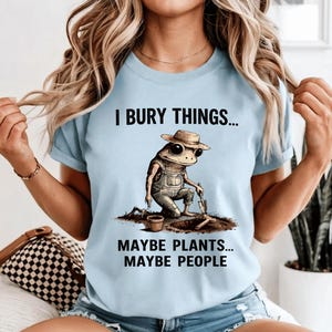Pode incluir: Camiseta azul clara com o texto "I BURY THINGS... MAYBE PLANTS... MAYBE PEOPLE." A camiseta apresenta um sapo de desenho animado usando macacão e chapéu, cavando na terra com uma pequena pá. Um pequeno vaso também está na imagem.