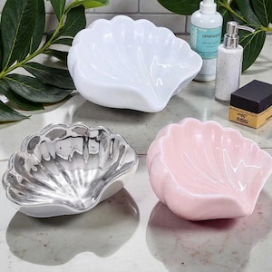 Puede incluir: Tres cuencos de cerámica con forma de concha en blanco, plateado y rosa. Los cuencos tienen un acabado brillante y están dispuestos sobre una superficie reflectante. El cuenco blanco está en la parte superior central, el plateado en la inferior izquierda y el rosa en la inferior derecha.