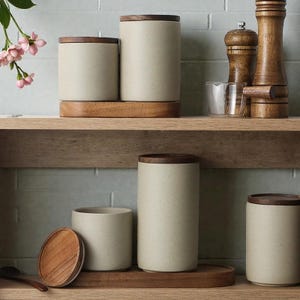 Puede incluir: Un conjunto de botes de cocina de cerámica beige con tapas de madera oscura. Los botes cilíndricos se exhiben en bandejas de madera. Un molinillo de pimienta de madera y otros artículos de cocina son visibles.