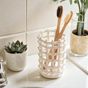 May include: A white ceramic toothbrush holder with a woven design, holding two wooden toothbrushes with black and tan bristles. A small succulent in a white pot and a glass vase with greenery are also visible.