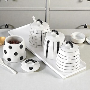 May include: A collection of white ceramic kitchen canisters and containers with black accents. The set includes jars with grid, stripe, and polka dot patterns, along with a small dish and a serving spoon. The canisters have black handles and lids.