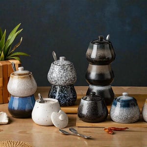 May include: A collection of ceramic spice jars with lids and small spoons. The jars are in various colors, including white, blue, and black, with speckled and glazed finishes. They are arranged on a wooden surface, suggesting a kitchen or dining setting.