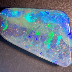 May include: A polished, trapezoidal-shaped opal gemstone featuring a vibrant play-of-colour with intense flashes of electric blue, bright emerald green, and subtle hints of orange and yellow set against a translucent, milky white base.