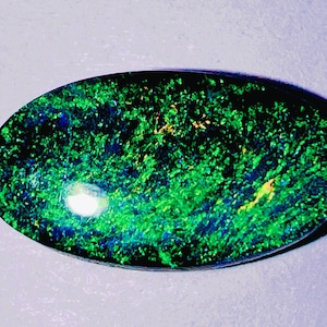 May include: An oval-shaped black opal gemstone featuring a dark base color with vibrant, scattered flashes of bright green and subtle hints of yellow and blue light reflecting across its polished, smooth, and slightly domed surface.