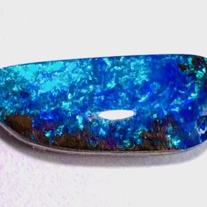 Op de afbeelding: Een gepolijste, langwerpige boulder-opaal edelsteen met een levendig kleurenspel met intense tinten elektrisch blauw, cyaan en diep marineblauw, gecontrasteerd door donkere, aards bruine ijzersteenmatrix-insluitingen langs de onderrand.