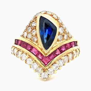 Pode incluir: Um anel de ouro com uma safira azul central em forma de escudo, rodeada por uma fila em V de rubis de corte quadrado e uma borda de diamantes redondos de corte brilhante. O design é simétrico com uma base pontiaguda e engaste em ouro.