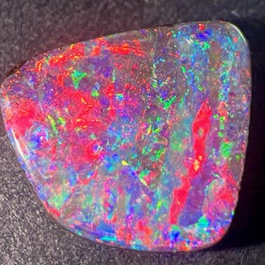May include: A polished, triangular-shaped opal gemstone featuring a dark base color with vibrant, multi-colored flashes of red, green, and blue light across its surface. The stone displays a high-gloss finish with a distinct play-of-color effect.