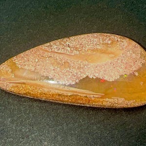 Op de afbeelding: Een traanvormige, gepolijste edelsteen met een mix van aardse bruine en beige tinten en een textuur die er zanderig uitziet. De steen vertoont doorschijnende amberkleurige secties met kleine, verspreide vlekjes in iriserende kleuren.