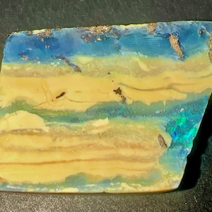Op de afbeelding: Een gepolijste, rechthoekige opaal edelsteen met horizontale banden van crème, beige en lichtbruin, gelaagd met doorschijnende blauwe en groene secties. Het oppervlak vertoont iriserende flitsen van helder blauw en groen licht.