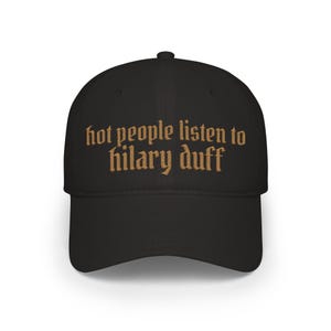 May include: Black baseball cap with the text "hot people listen to hilary duff" in a brown, gothic-style font. The cap has a curved brim and a structured crown, suitable for casual wear.