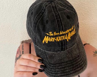 May include: A black, acid-washed baseball cap featuring the yellow embroidered text The New Adventures of MARY-KATE & ASHLEY on the front panel. The cap has a curved brim, a six-panel construction with stitched eyelets, and a button on the top.