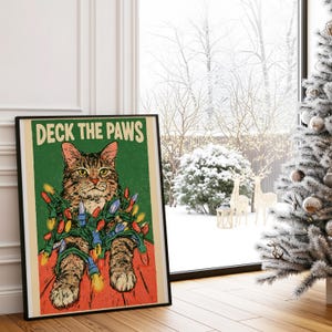 Könnte beinhalten: Ein rechteckiger Wandkunstdruck mit einer braunen getigerten Katze, die in bunte Weihnachtsbeleuchtung gewickelt ist, vor einem grünen Hintergrund. Der Text DECK THE PAWS steht in großen weißen Buchstaben oben. Die Katze liegt auf einer roten Fläche.