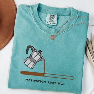May include: A turquoise Comfort Colors t-shirt with a coffee maker graphic and the text "MOTIVATION LOADING..". A gold necklace with a pendant is draped across the shirt. The shirt is folded.