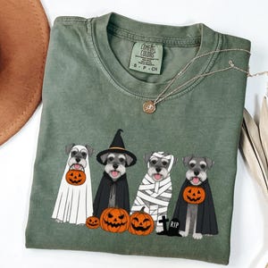 Puede incluir: Una camiseta verde oliva con un gráfico de cuatro perros Schnauzer disfrazados de Halloween, incluyendo un fantasma, una bruja, una momia y un vampiro con capas y cubos de calabaza, junto a tres calabazas de color naranja.