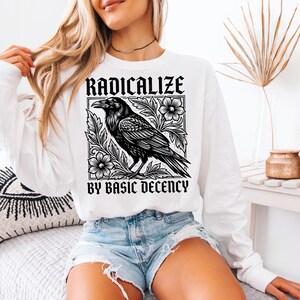 May include: A white long-sleeve sweatshirt featuring a black graphic print of a raven surrounded by floral elements. The text RADICALIZE is printed above the bird, and the text BY BASIC DECENCY is printed below the bird in a bold font.