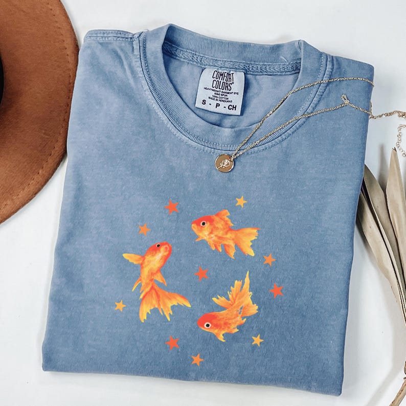 May include: A blue short-sleeved t-shirt with a Comfort Colors label in the collar. The shirt features a graphic print of three orange goldfish swimming in a circle, surrounded by small orange stars. A gold-toned necklace rests on the neckline.
